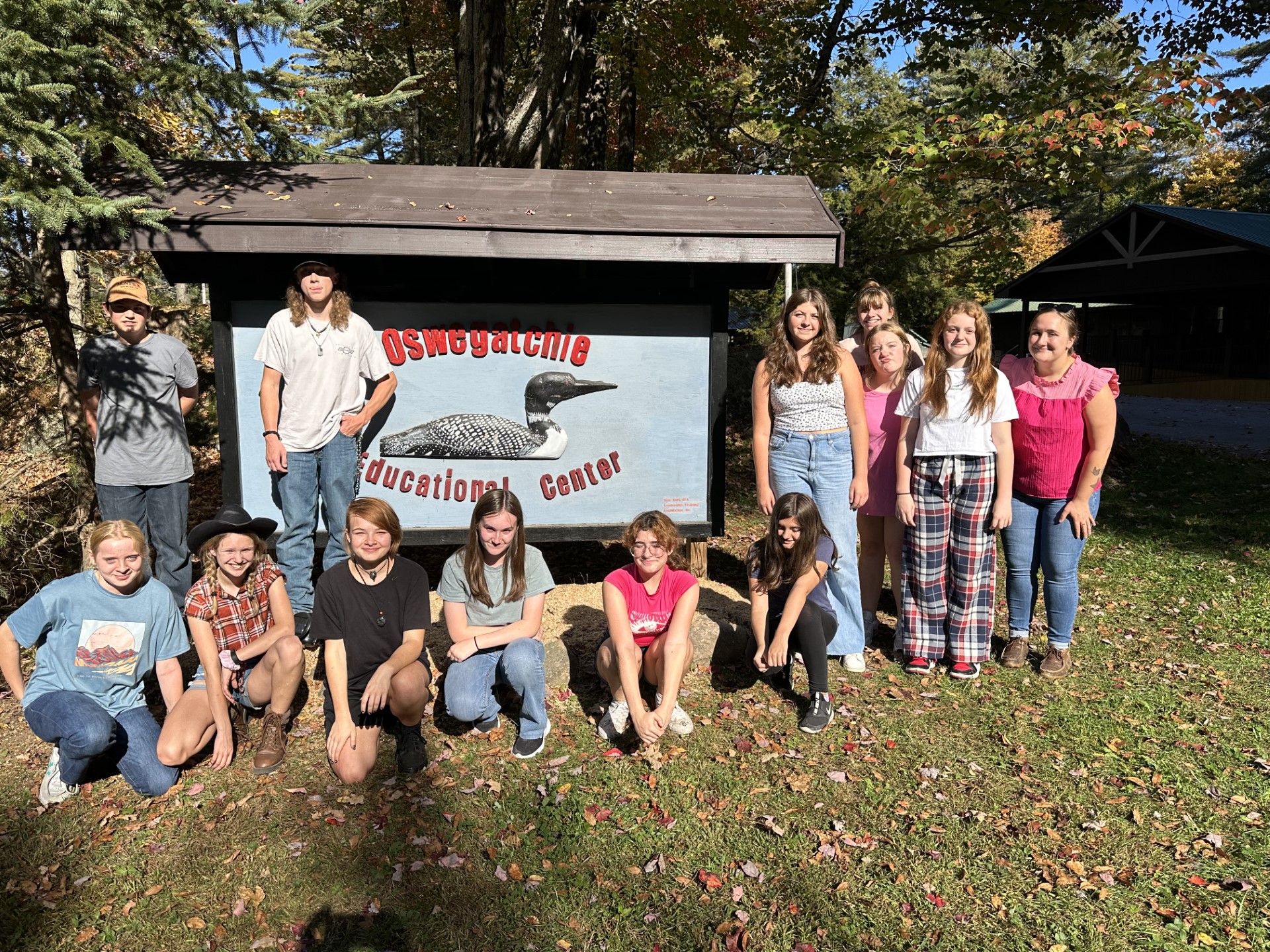 13 students stand or sit in front of the Camp Oswegatchie sign