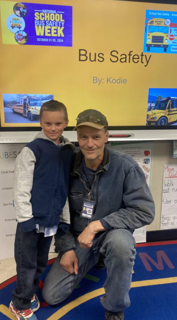 A students stands with a bus driver who is kneeling in front of a board about bus safety.